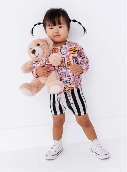 A toddler girl with black pigtails stands against a white studio background, holding a light brown teddy bear. She is wearing a pink long-sleeve top with a multicolor graphic print, black and white vertically striped shorts, and white low-top sneakers.