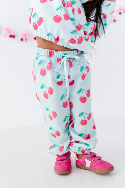 A child wearing a light blue cherry print sweatshirt and matching jogger pants with pink sneakers, shown from the waist down against a white background.