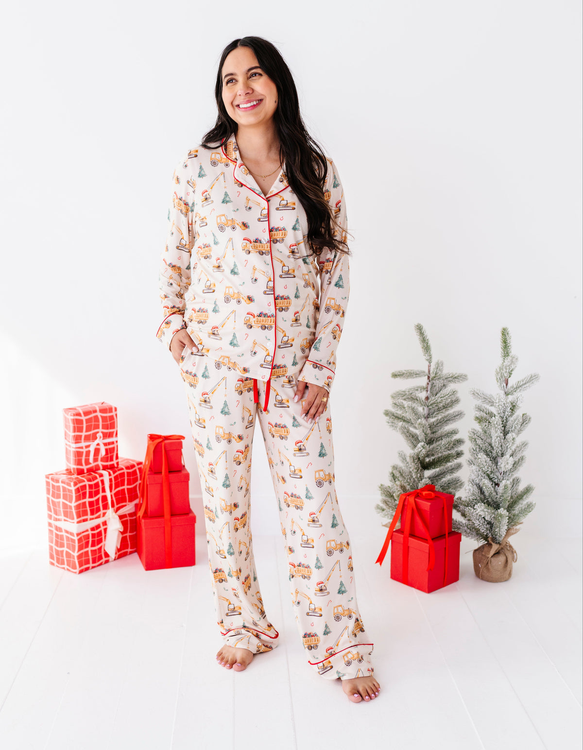 A woman wearing a festive Christmas-themed bamboo pajama set stands in a bright white room, flanked by wrapped holiday gifts and small artificial trees.