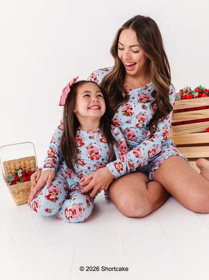 Woman and child wearing matching Strawberry Shortcake™ bamboo pajamas sitting on a white background with a wooden crate and basket of strawberries.