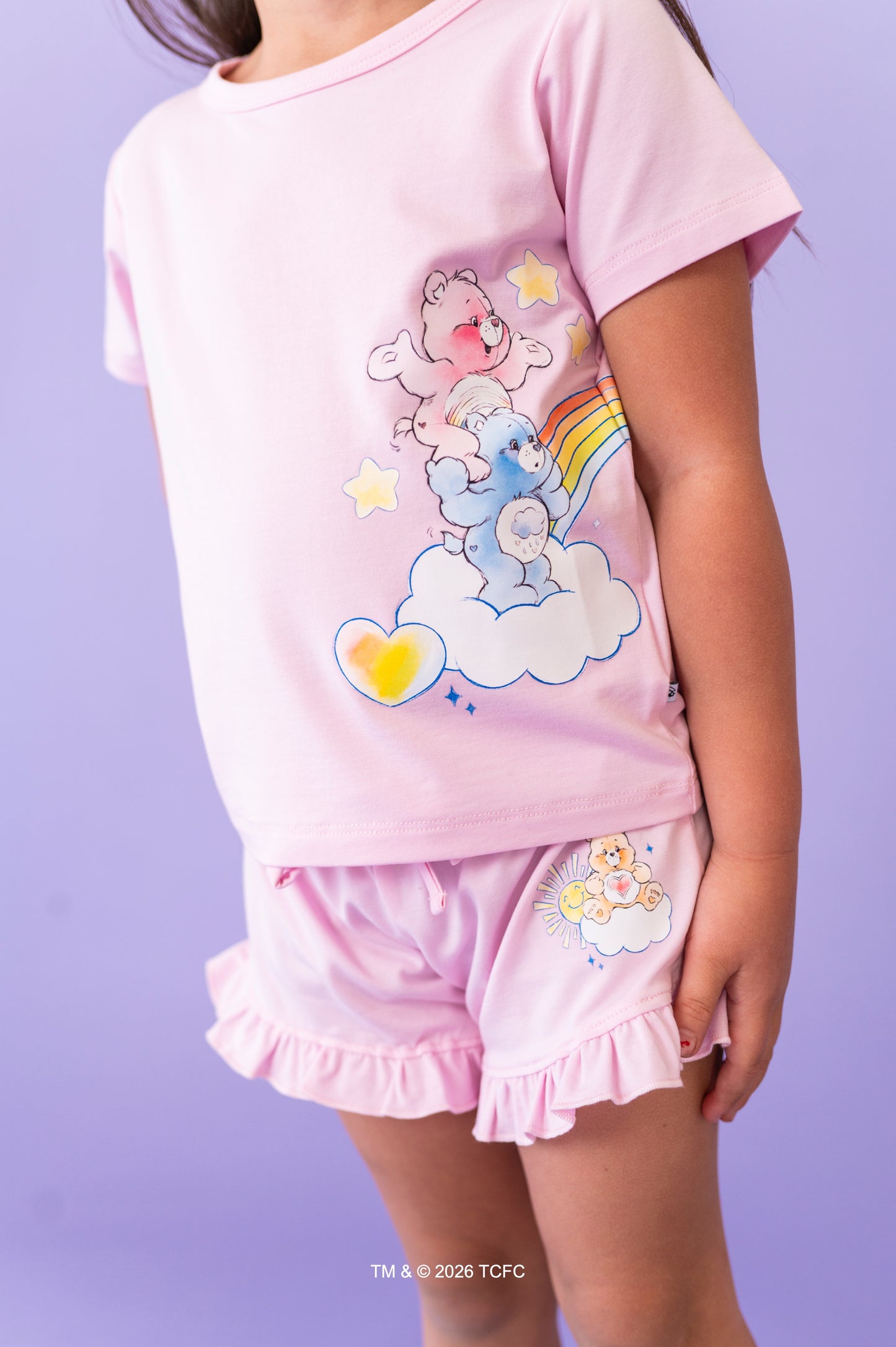 Child wearing a lilac short set with cartoon Care Bears™ design on a purple background