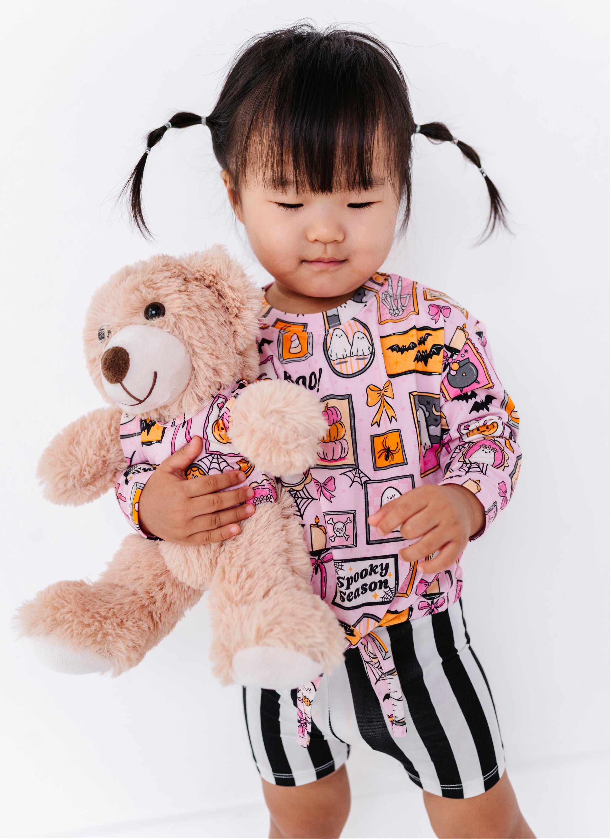 A toddler girl with black pigtails stands against a white background, holding a plush teddy bear. She is wearing a pink long-sleeve top with a colorful cartoon patchwork print and black-and-white striped shorts.