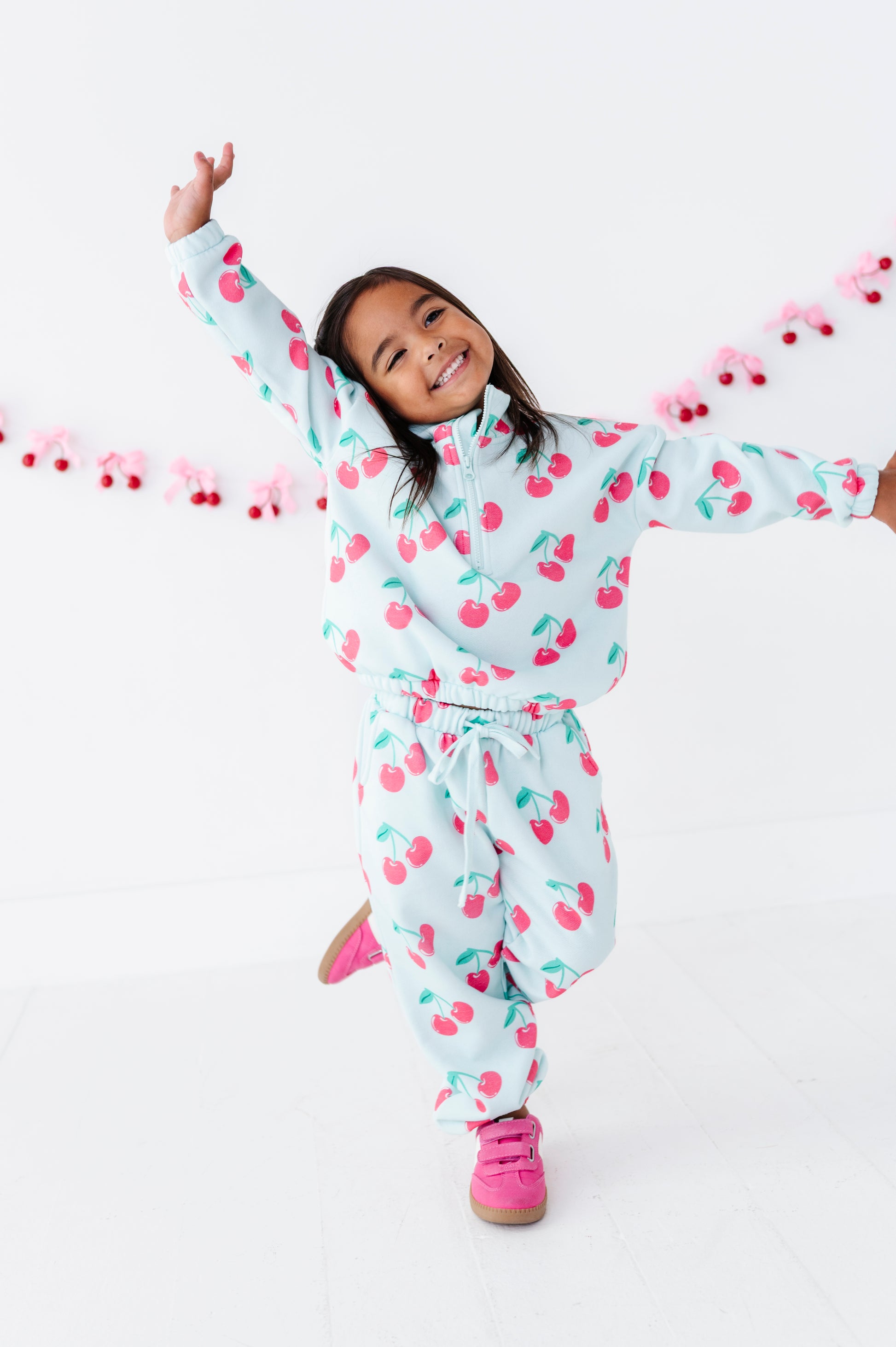 A young girl smiling and posing in  cherry-patterned zip-up sweatshirt  and matching sweatpants, with bright pink sneakers, against a clean white studio background with a pink cherry garland.