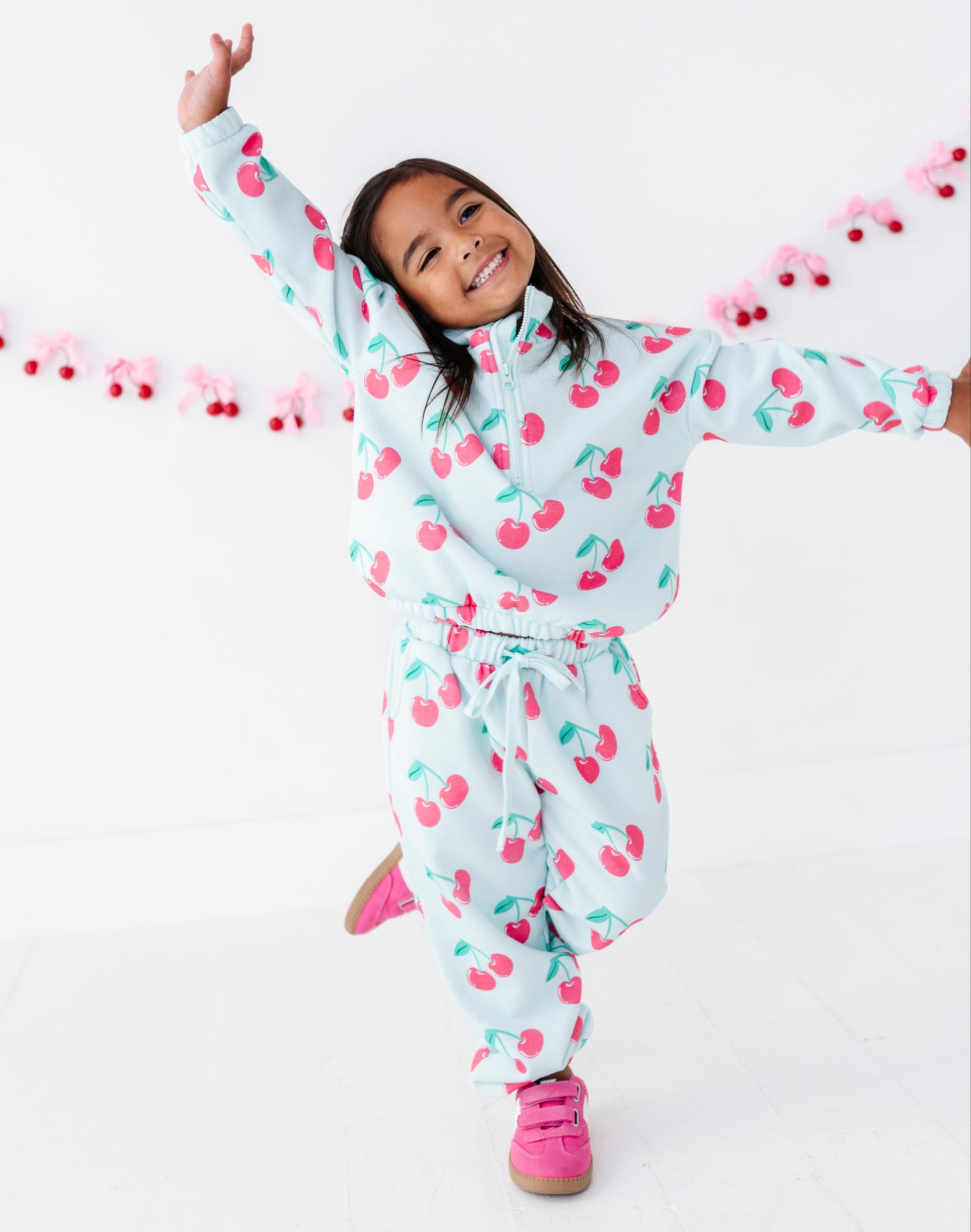 A young girl smiling and posing in a white cherry-patterned zip-up hoodie and matching sweatpants, with bright pink sneakers, against a clean white studio background with a pink cherry garland.