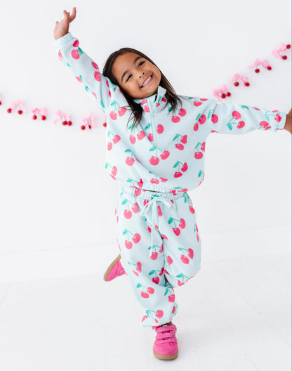 A young girl smiling and posing in a white cherry-patterned zip-up hoodie and matching sweatpants, with bright pink sneakers, against a clean white studio background with a pink cherry garland.
