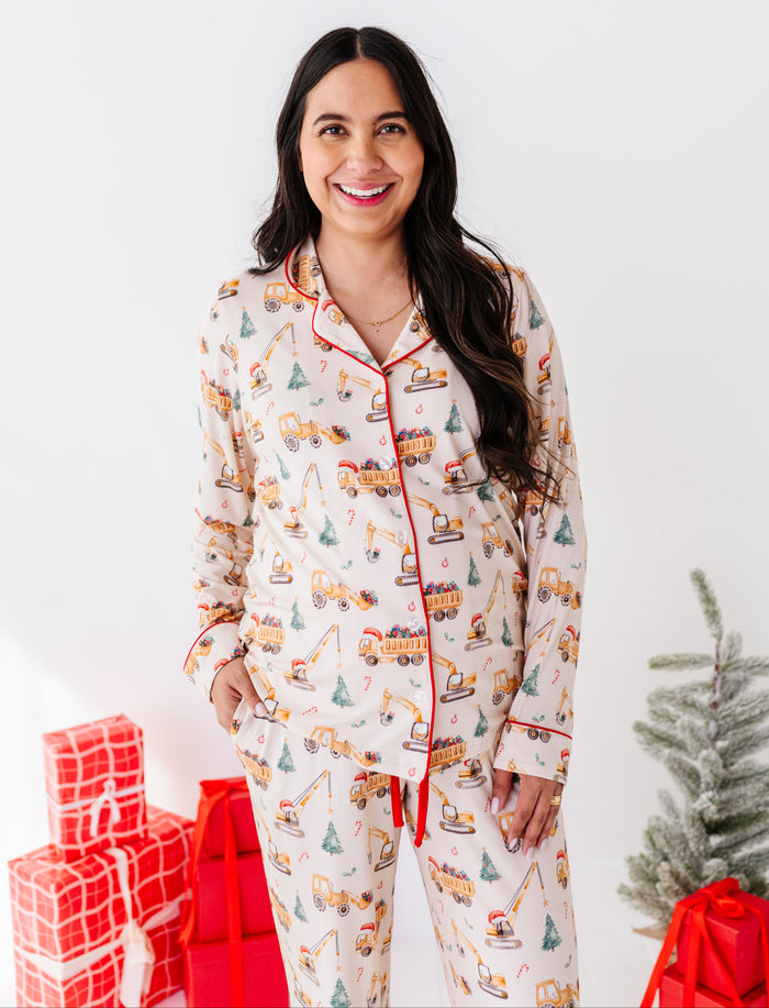 A woman wearing a matching pajama set with a construction truck pattern, standing in front of a white wall with Christmas decorations including wrapped gifts and a small tree.