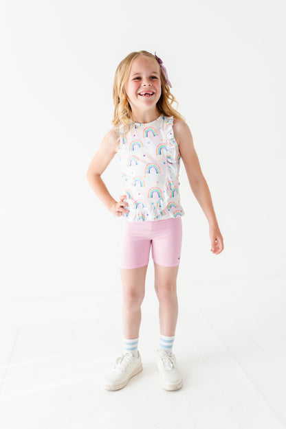 Young girl wearing a sleeveless bamboo cotton peplum top with watercolor rainbow pattern and pink bamboo biker shorts on a white background