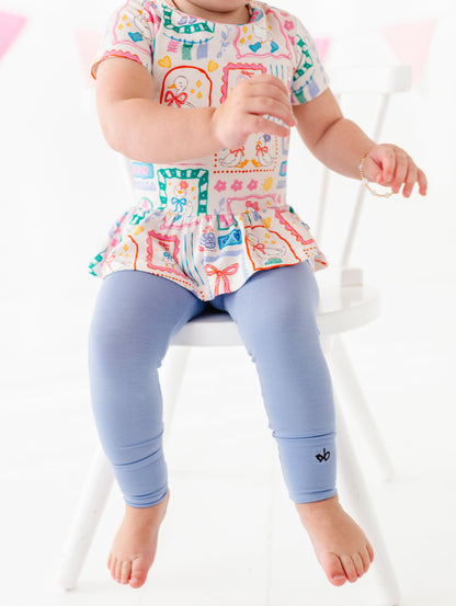 Child wearing a colorful top and blue leggings sitting on a white chair.