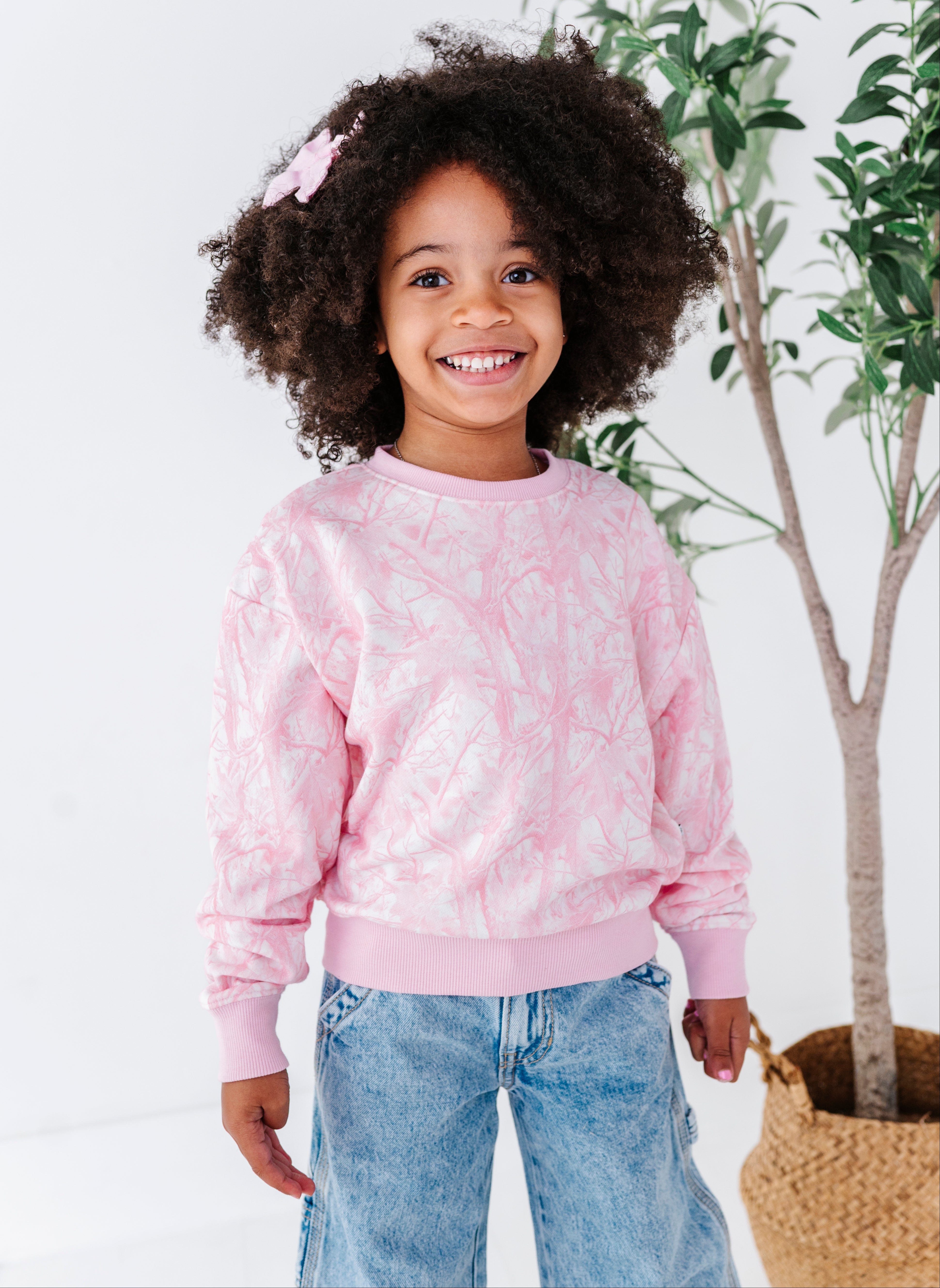 Child wearing a pink camo sweatshirt and blue jeans standing in front of a plant.