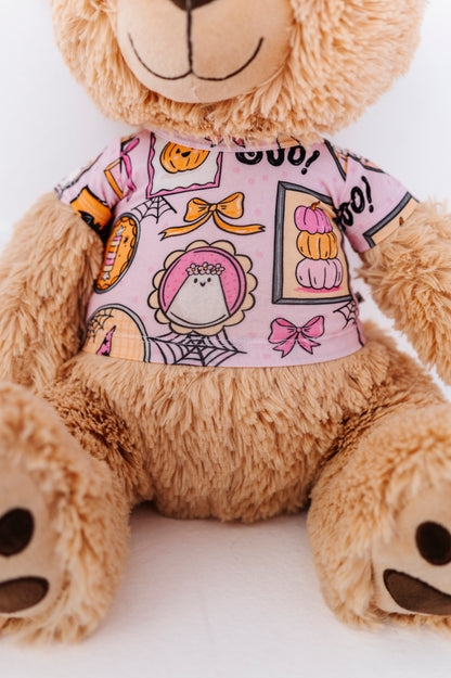 A close-up of a plush teddy bear wearing a pink short-sleeve shirt with a Halloween-themed print featuring pumpkins, ghosts, spiderwebs, and bows.