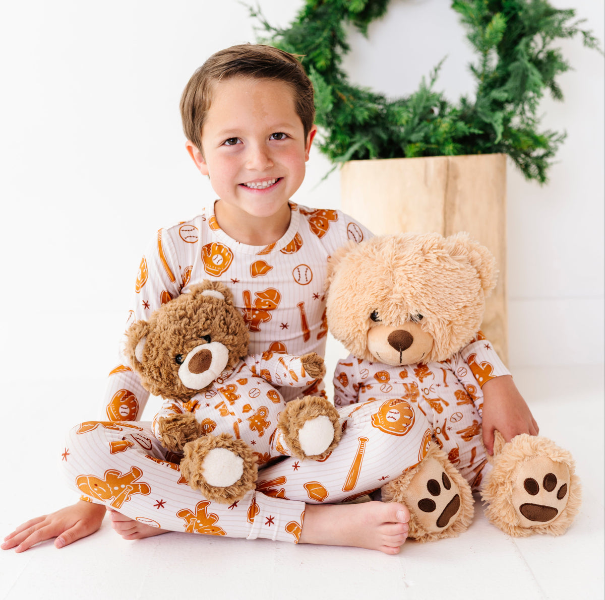 A young boy wearing a white and orange pumpkin-themed pajama set sits cross-legged on a white floor, holding two teddy bears. A green wreath in a wooden box is in the background.