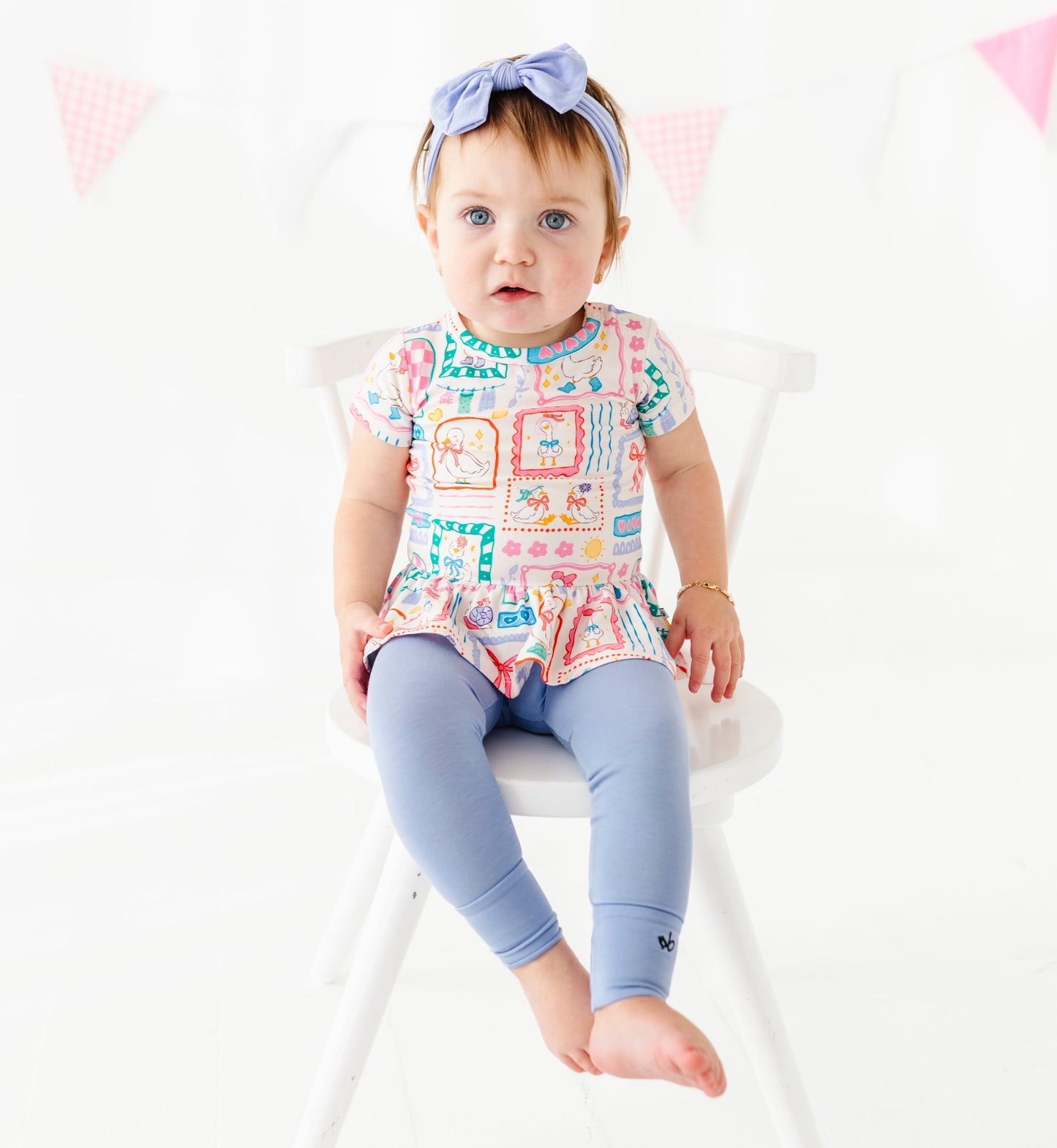 Baby sitting on a chair wearing a colorful patterned shirt and blue leggings with a white background.