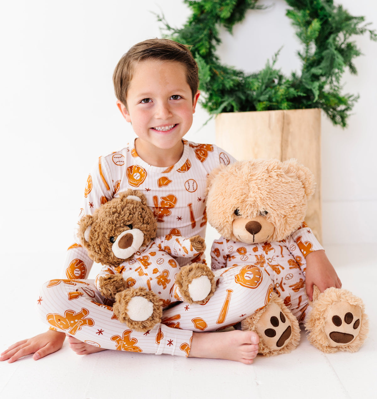 A young boy wearing a white and orange pumpkin-themed pajama set sits cross-legged on a white floor, holding two teddy bears. A green wreath in a wooden box is in the background.