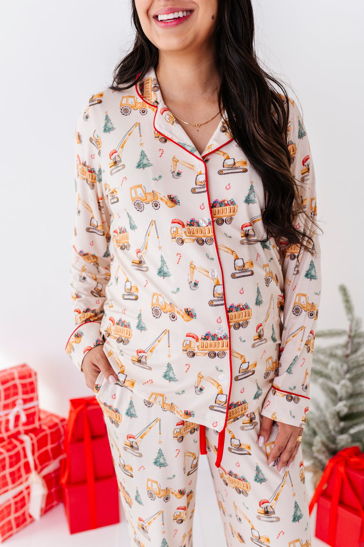 A woman wearing a long-sleeve bamboo pajama set with an all-over construction truck print and red piping trim, standing in a festive holiday setting with wrapped gifts and a Christmas tree.