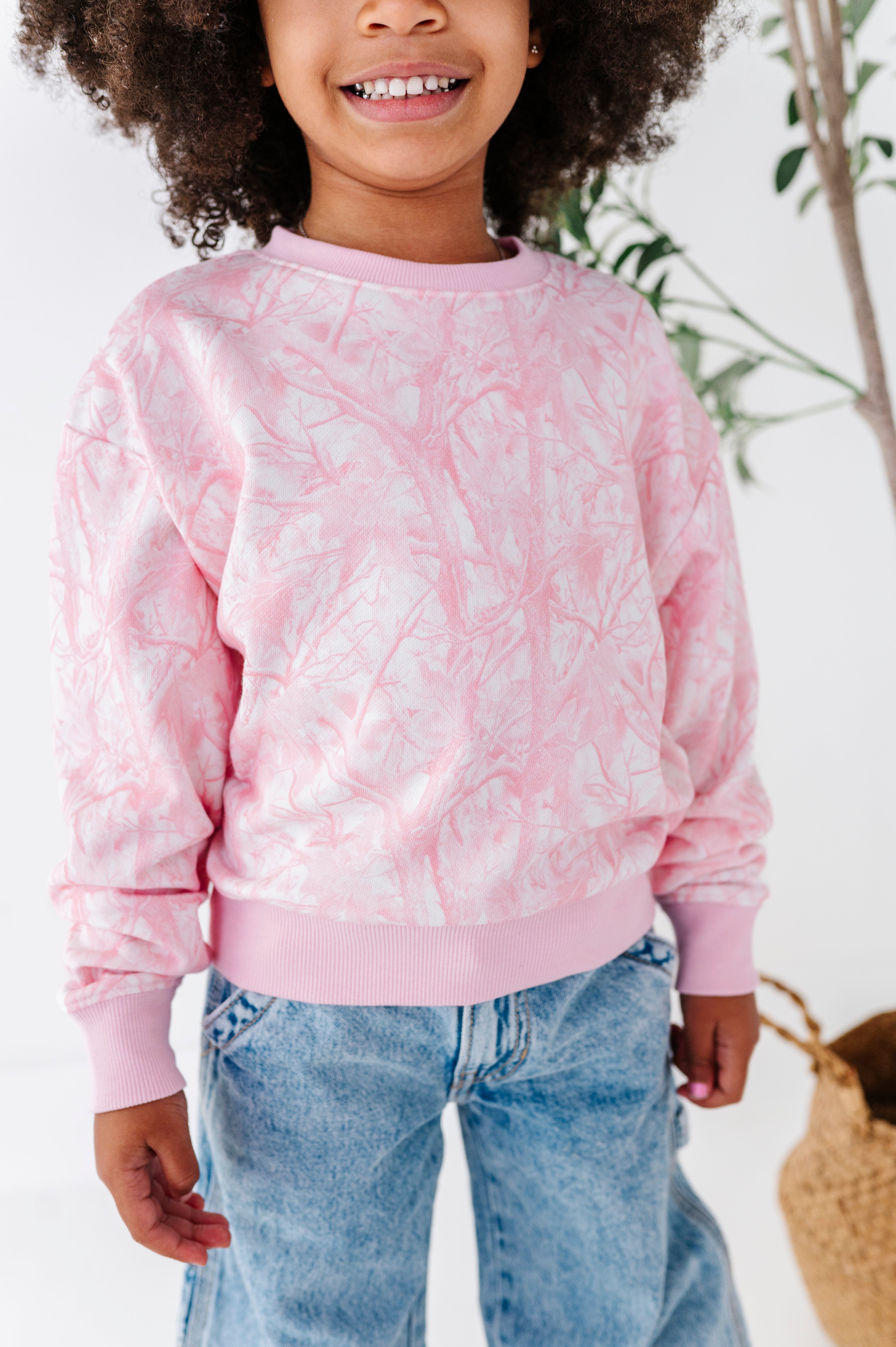 Child wearing a pink tree camo sweatshirt and blue jeans with a blurred background