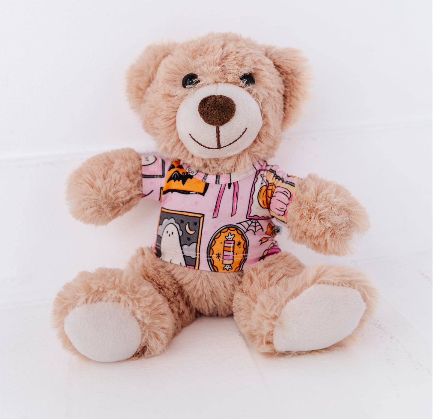 A plush teddy bear in light brown faux fur wearing a pink graphic t-shirt, sitting against a seamless white background.