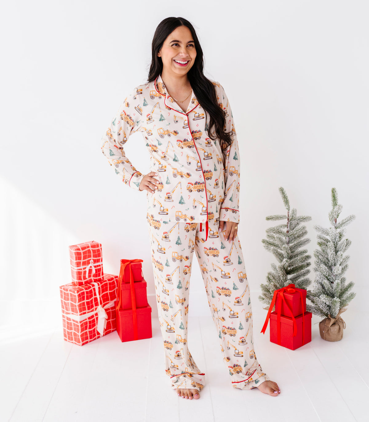 A woman stands barefoot in a bright white room wearing a long-sleeve Christmas construction-print pajama set with a holiday village print. She is flanked by red wrapped gift boxes and two small artificial Christmas trees.