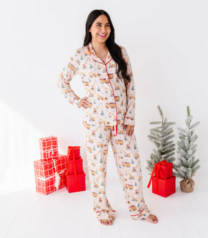A woman stands barefoot in a bright white room wearing a long-sleeve Christmas construction-print pajama set with a holiday village print. She is flanked by red wrapped gift boxes and two small artificial Christmas trees.