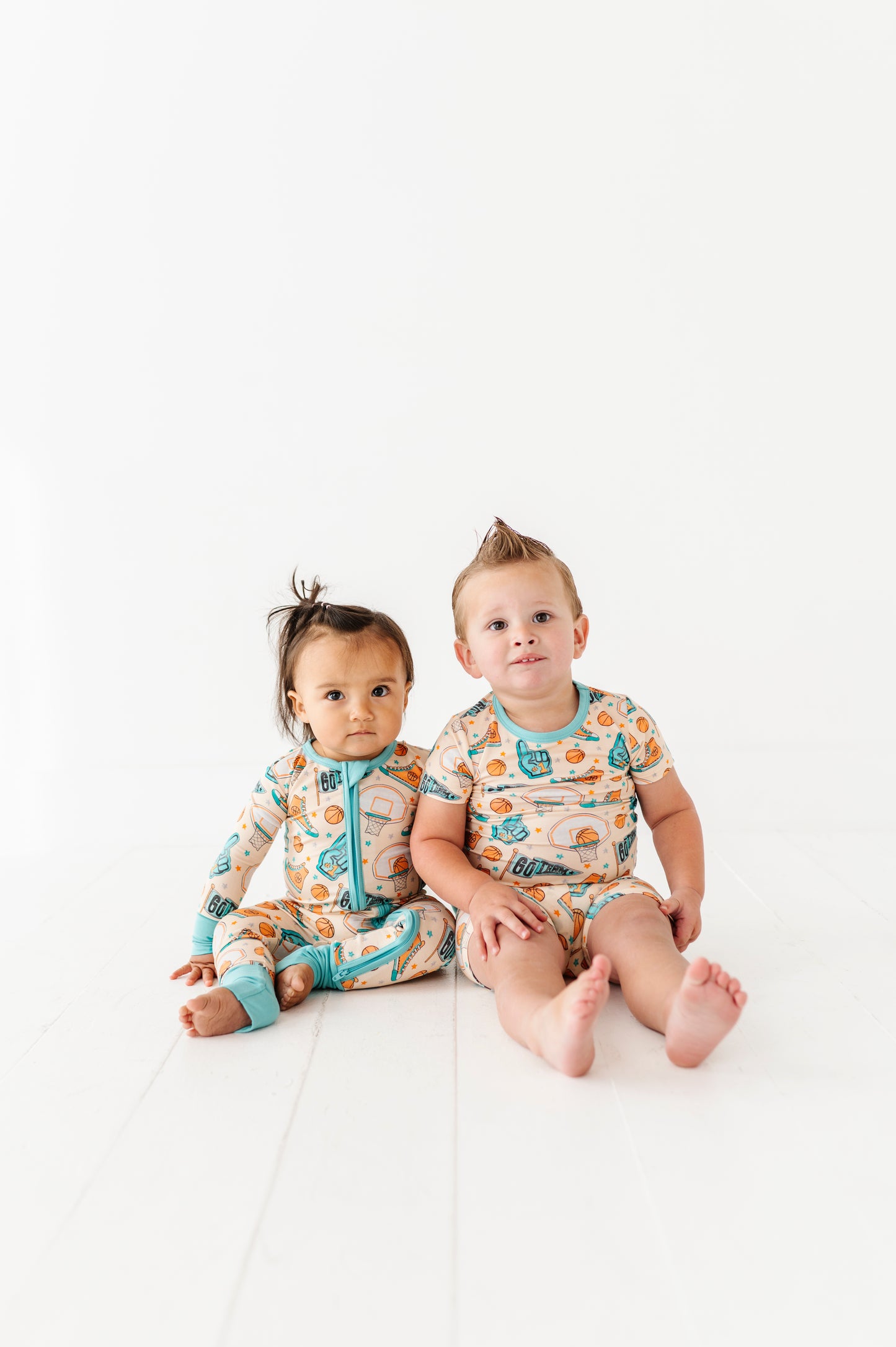 Two children wearing sports themed bamboo onesies sitting on a white surface.