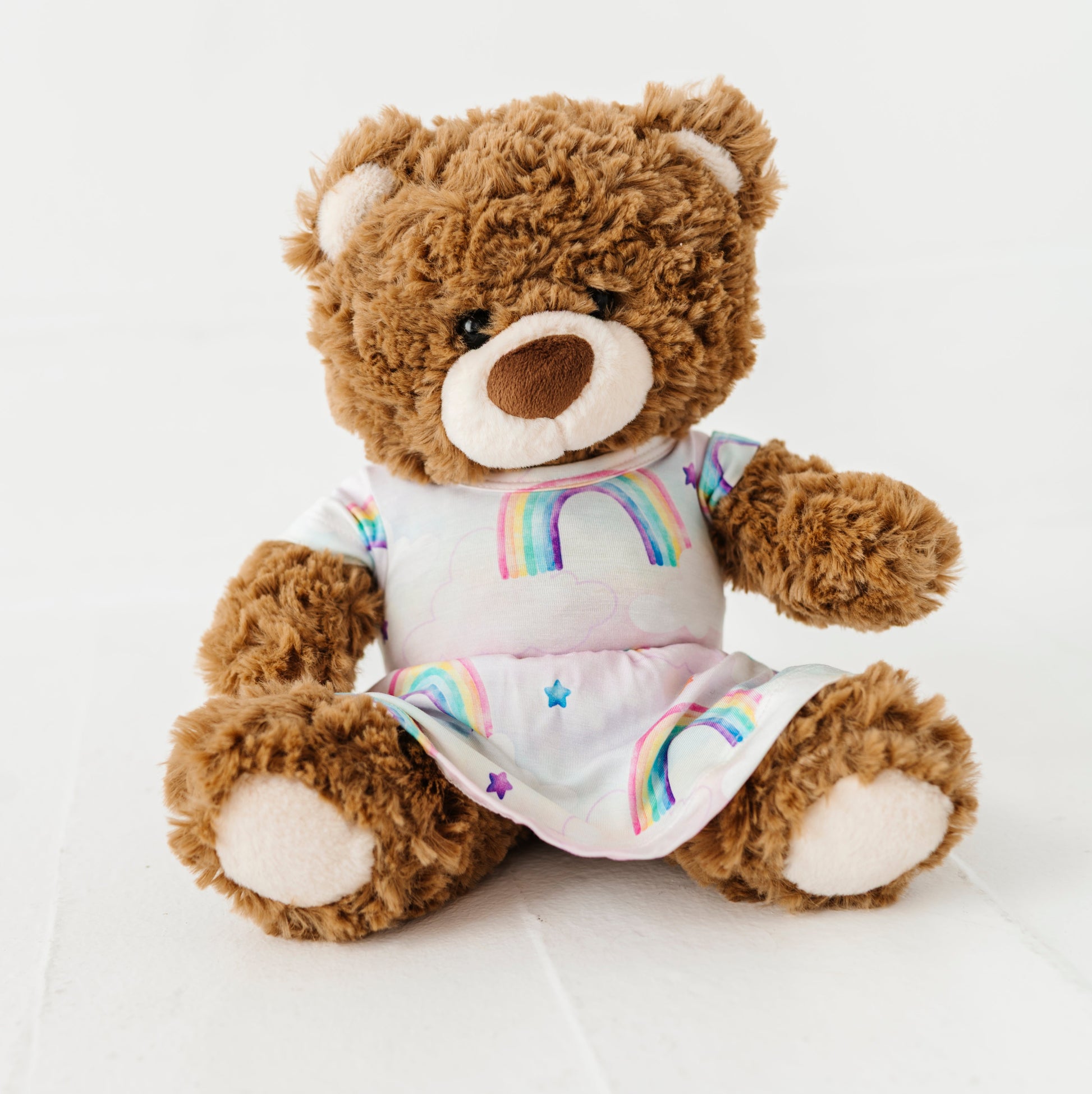 Brown teddy bear wearing a dress with rainbow designs on a light gray background