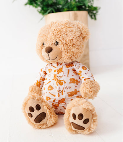 A plush teddy bear sitting upright, wearing a white long-sleeve pajama top with an orange and yellow gingerbread cookie pattern. The bear has light brown fur and is posed in front of a light-colored wooden box with a green wreath on top, set against a bright white background.