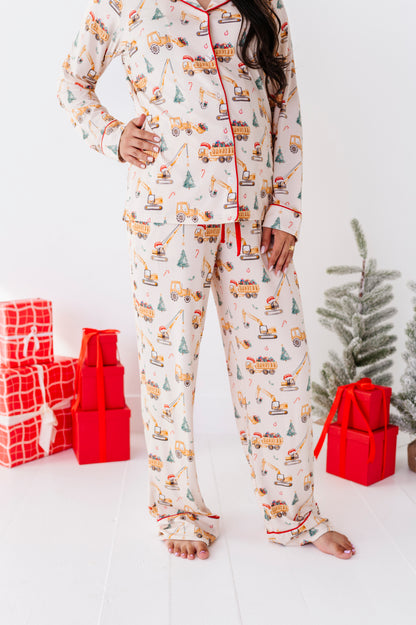 A woman wearing a two-piece bamboo pajama set with an all-over construction truck print and red piping, standing barefoot in a bright room with wrapped gifts and a small tree.