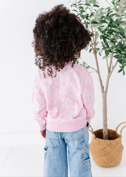 Child wearing a pink sweatshirt with pink camo pattern, standing in front of a plant against a white wall.