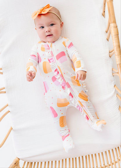 A baby wearing a footed sleep sack with a pink, orange, and yellow abstract pattern, lying on a white muslin mattress in a rattan bassinet. The baby is also wearing an orange bow headband.