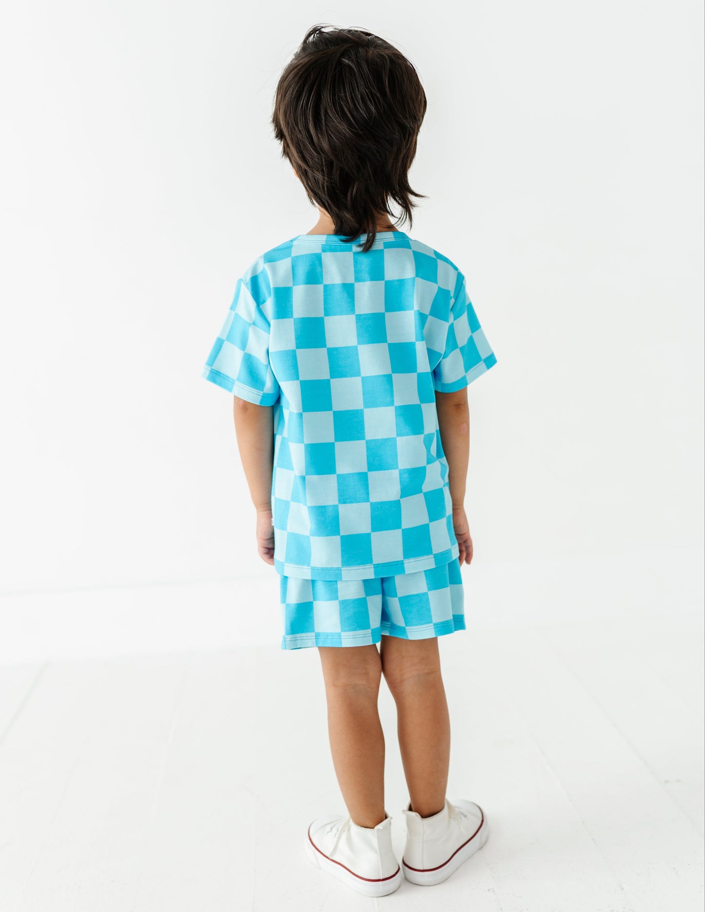 A child standing with their back to the camera, wearing a matching light blue and white checkered short-sleeve t-shirt and shorts set, paired with white sneakers.