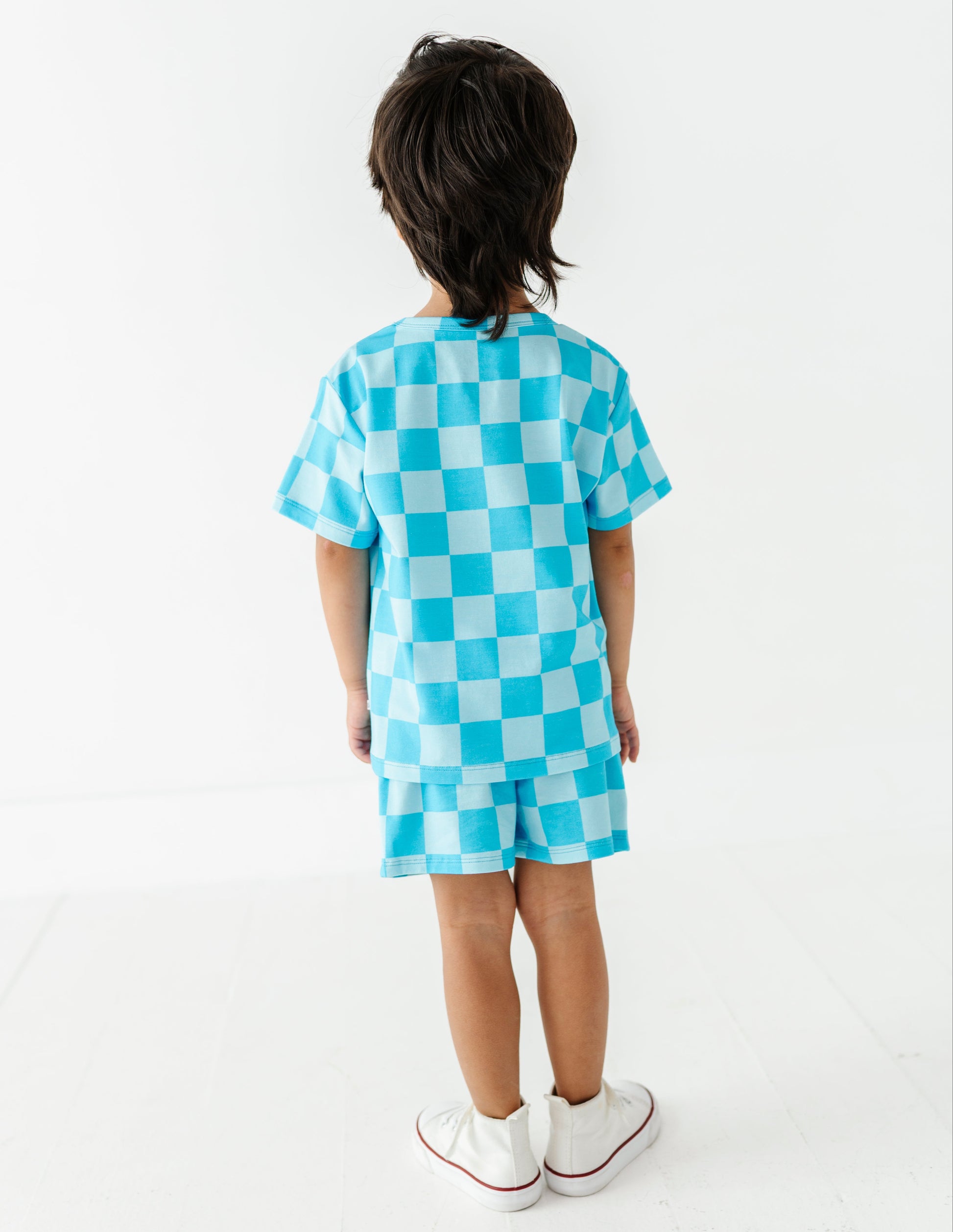 A child standing with their back to the camera, wearing a matching light blue and white checkered short-sleeve t-shirt and shorts set, paired with white sneakers.