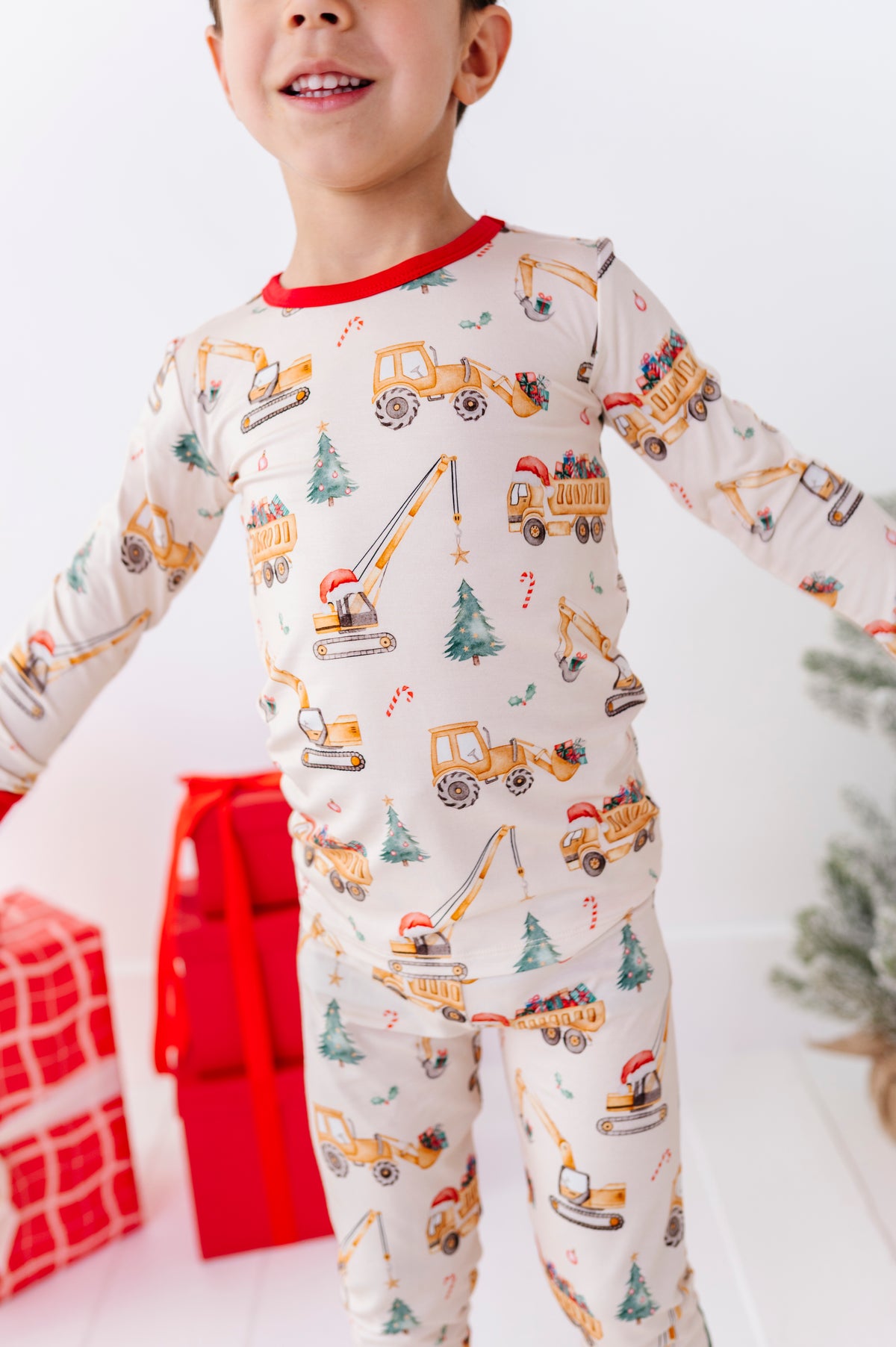 A child wearing a long-sleeve bamboo pajama set with an all-over construction vehicle print, featuring yellow excavators, dump trucks, and cranes on an off-white background with red trim. The child is standing in a festive setting with wrapped gifts and a small Christmas tree.