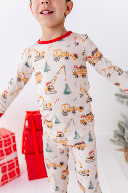 A child wearing a long-sleeve bamboo pajama set with an all-over construction vehicle print, featuring yellow excavators, dump trucks, and cranes on an off-white background with red trim. The child is standing in a festive setting with wrapped gifts and a small Christmas tree.