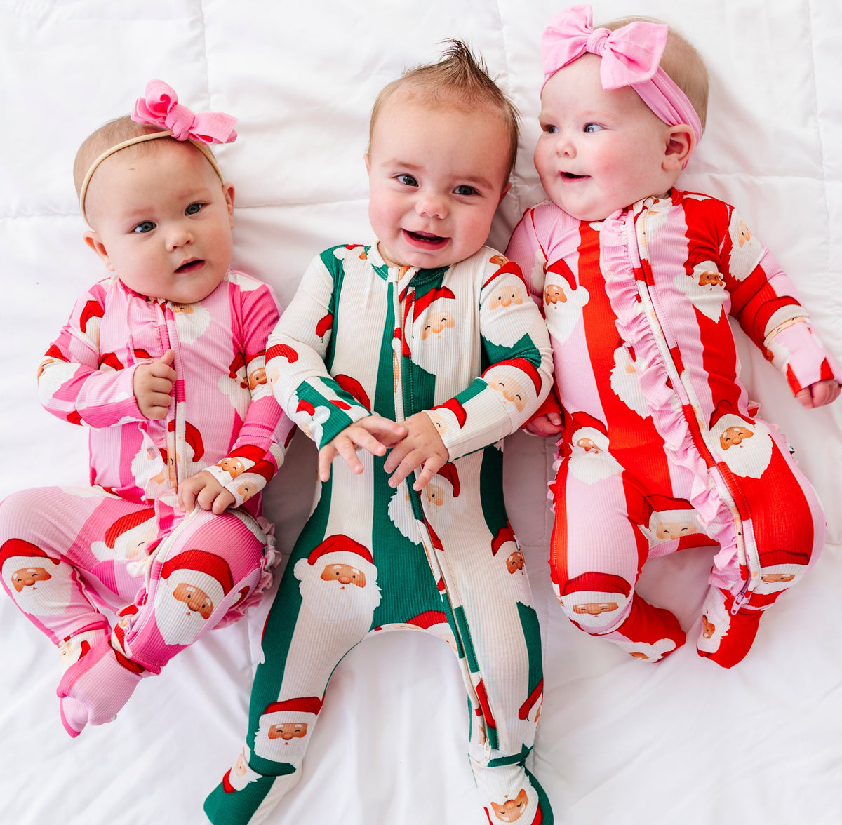 Three babies lying side by side on a white quilted blanket, wearing festive Santa Claus print footed pajamas. The baby on the left wears a pink pajama set with a pink bow headband.
