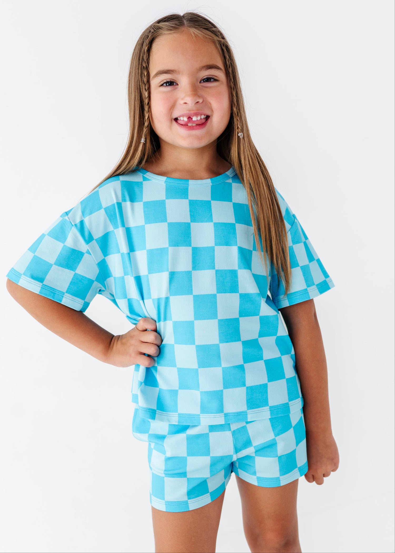 A young girl with long light brown hair smiling, wearing a matching short-sleeve t-shirt and shorts set in a light blue and white checkerboard pattern, set against a plain white studio background.