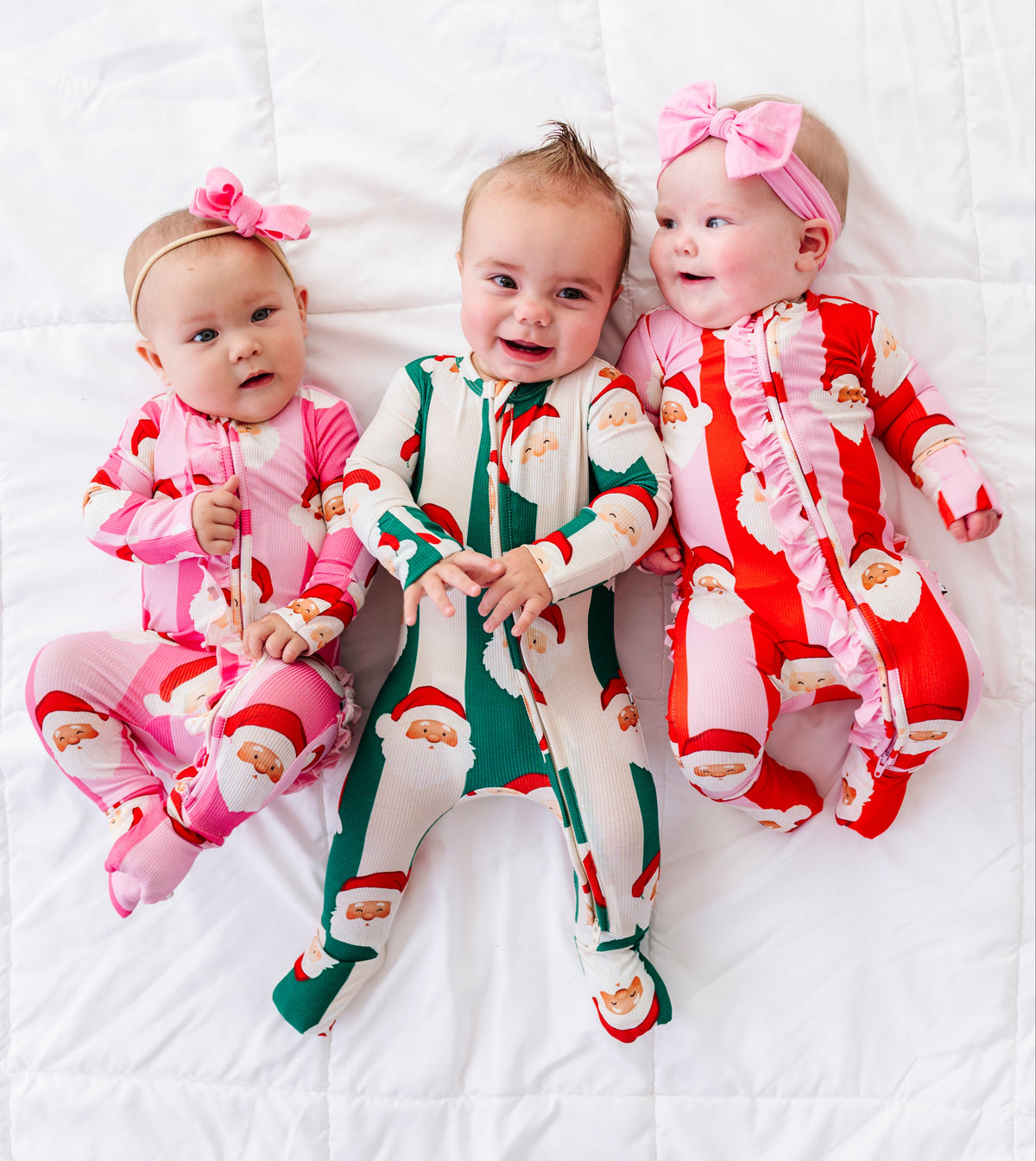 Three babies lying side by side on a white quilted blanket, wearing festive Santa Claus footed bamboo pajamas. The baby on the left wears a pink pajama set with a pink bow headband.