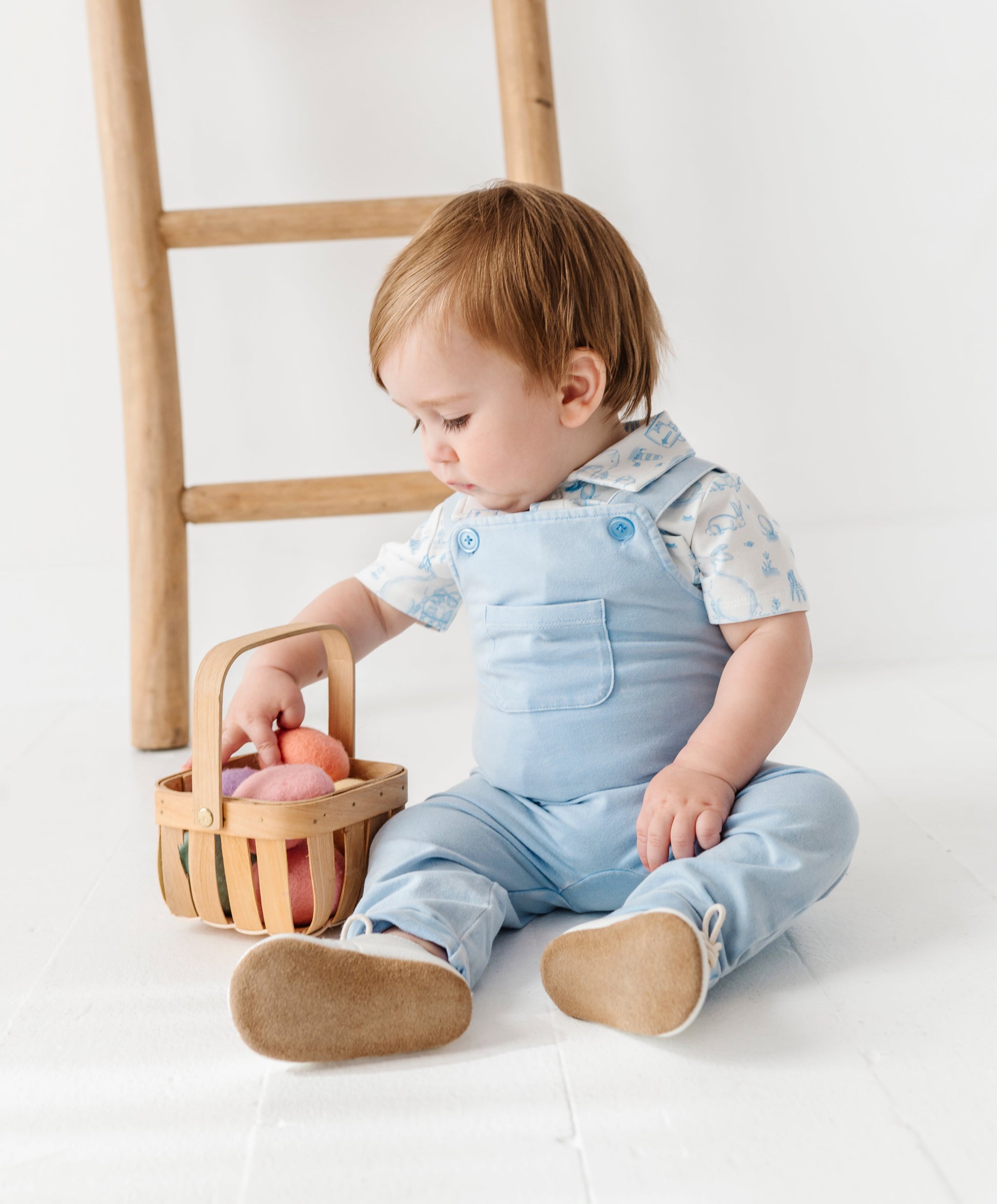 Baby in light blue overalls sitting on a white floor with a wooden basket.