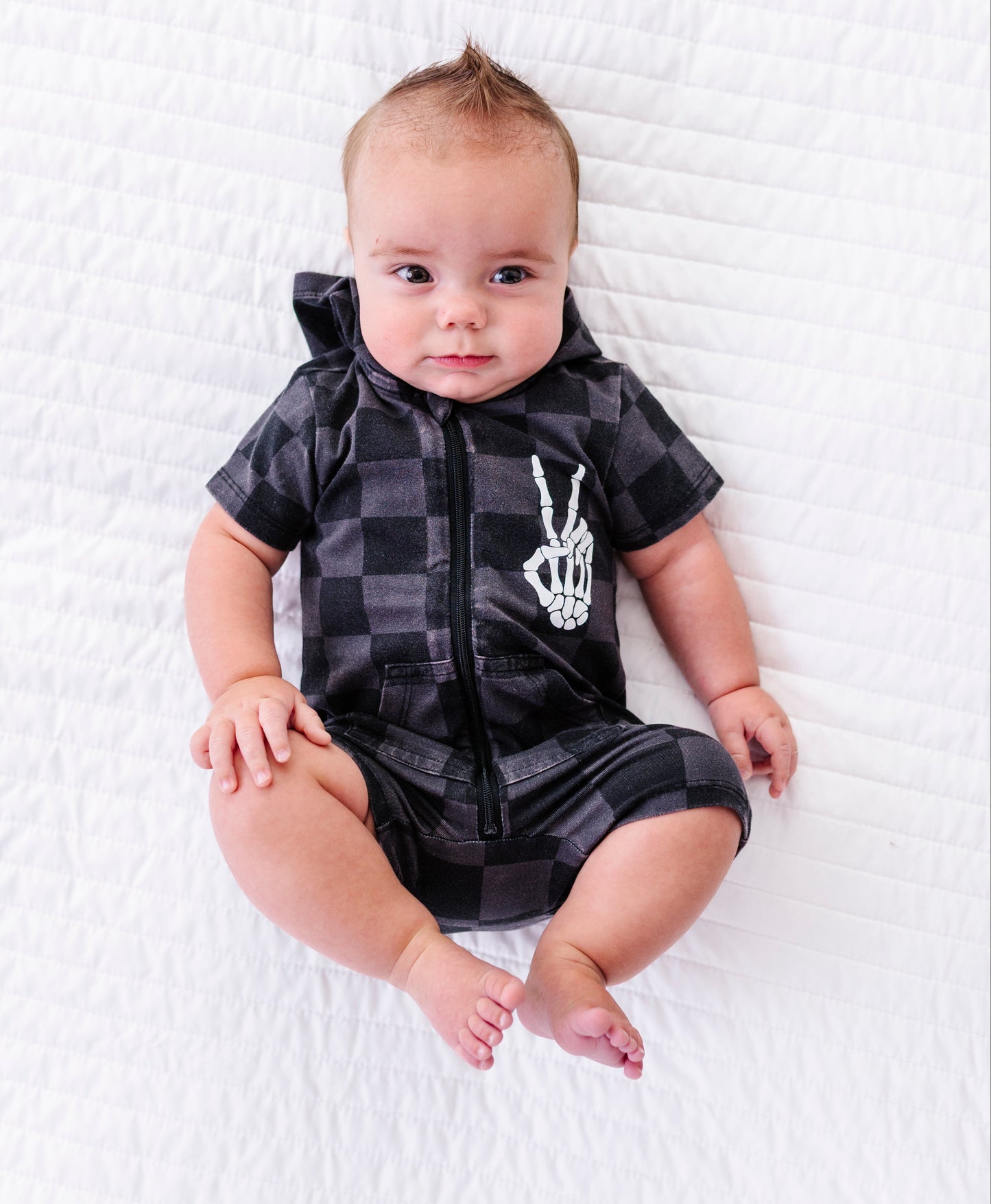 A baby lying on a white quilted blanket, wearing a black and gray plaid hooded romper with a white skeleton hand graphic on the chest.