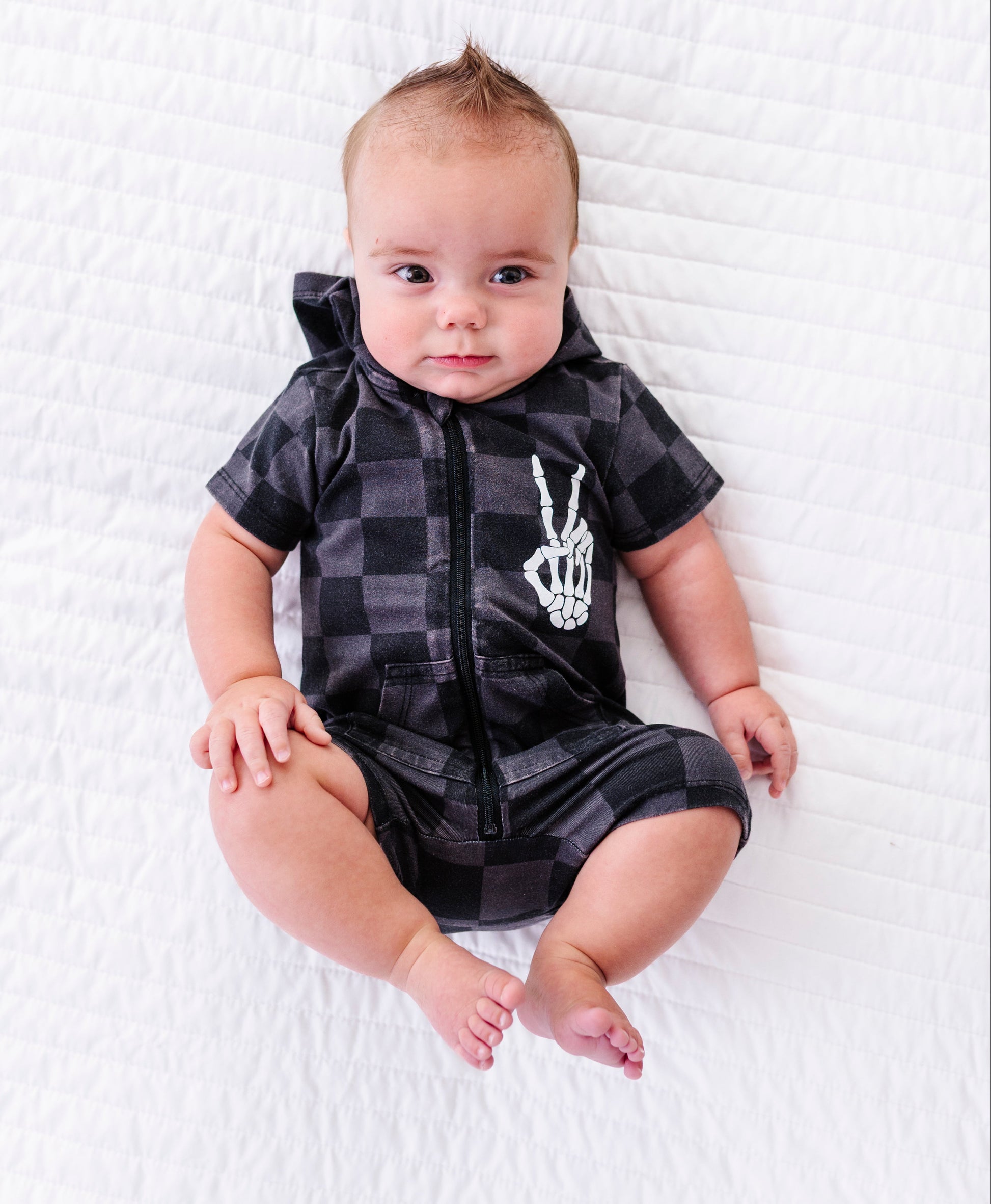 A baby lying on a white quilted blanket, wearing a black and gray plaid hooded romper with a white skeleton hand graphic on the chest.