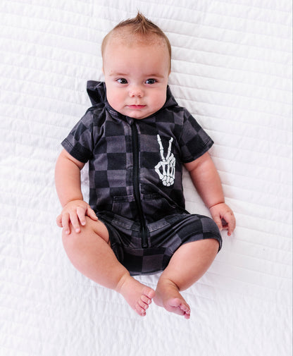 A baby lying on a white quilted blanket, wearing a black and gray plaid hooded romper with a white skeleton hand graphic on the chest.