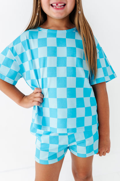 A young girl wearing a matching light blue and white checkerboard short-sleeve t-shirt and shorts set, shown from the chest down against a plain white background.