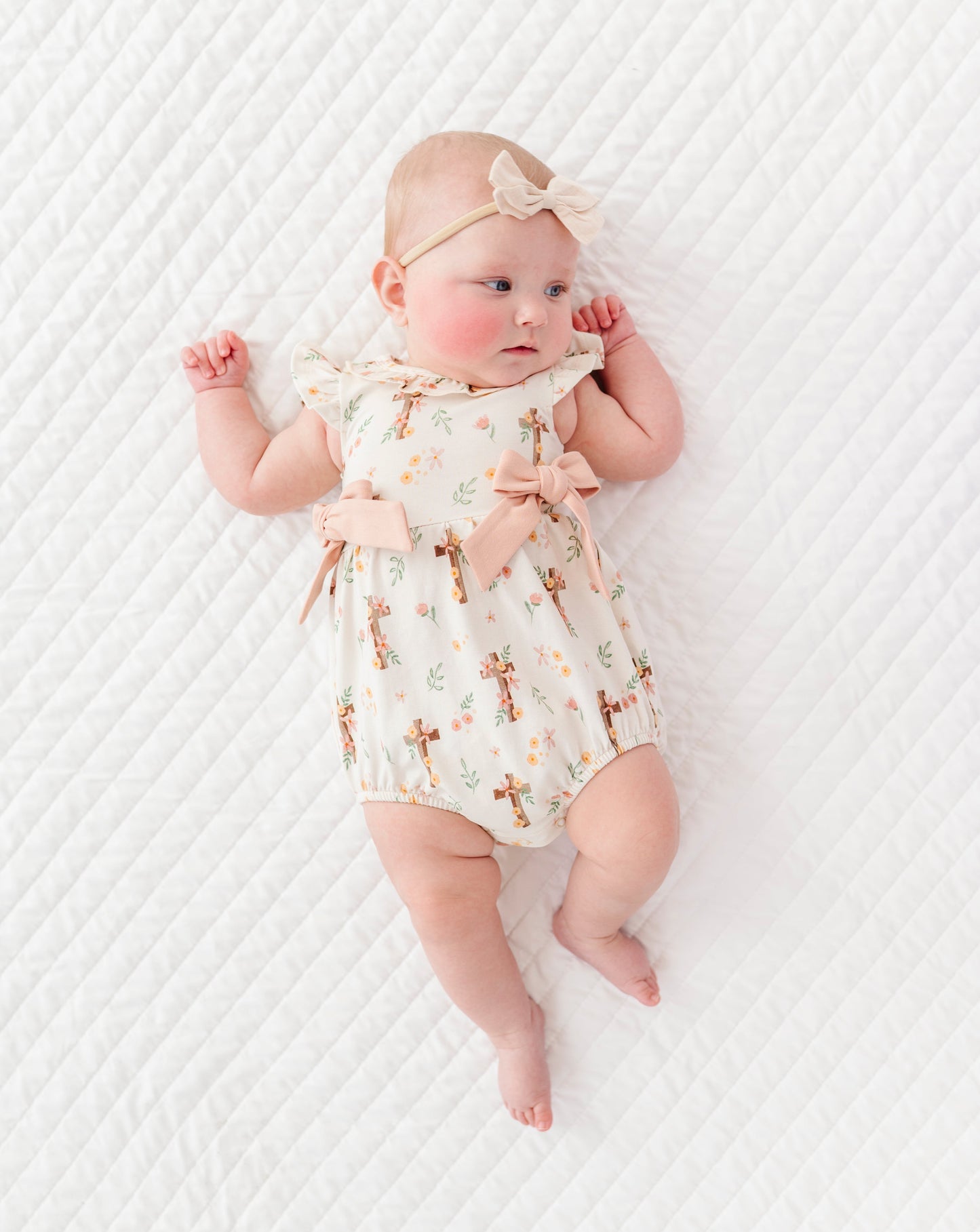 baby girl in a cross with florals bubble romper and a neutral colored headband on a white background 