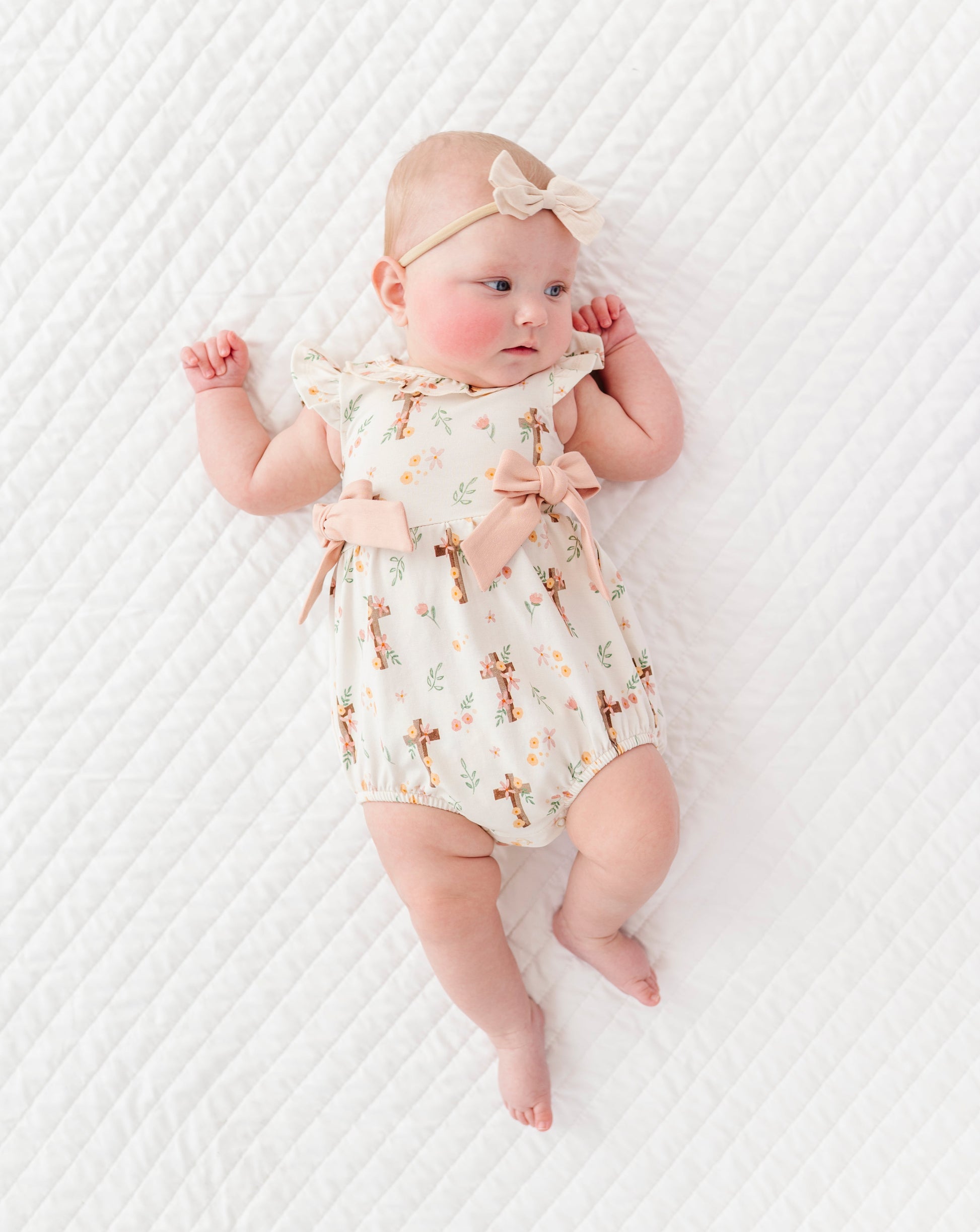 baby girl in a cross with florals bubble romper and a neutral colored headband on a white background 