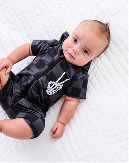A baby lying on a white quilted blanket, wearing a black and gray checkered short-sleeve romper with a white skeleton hand graphic on the chest.