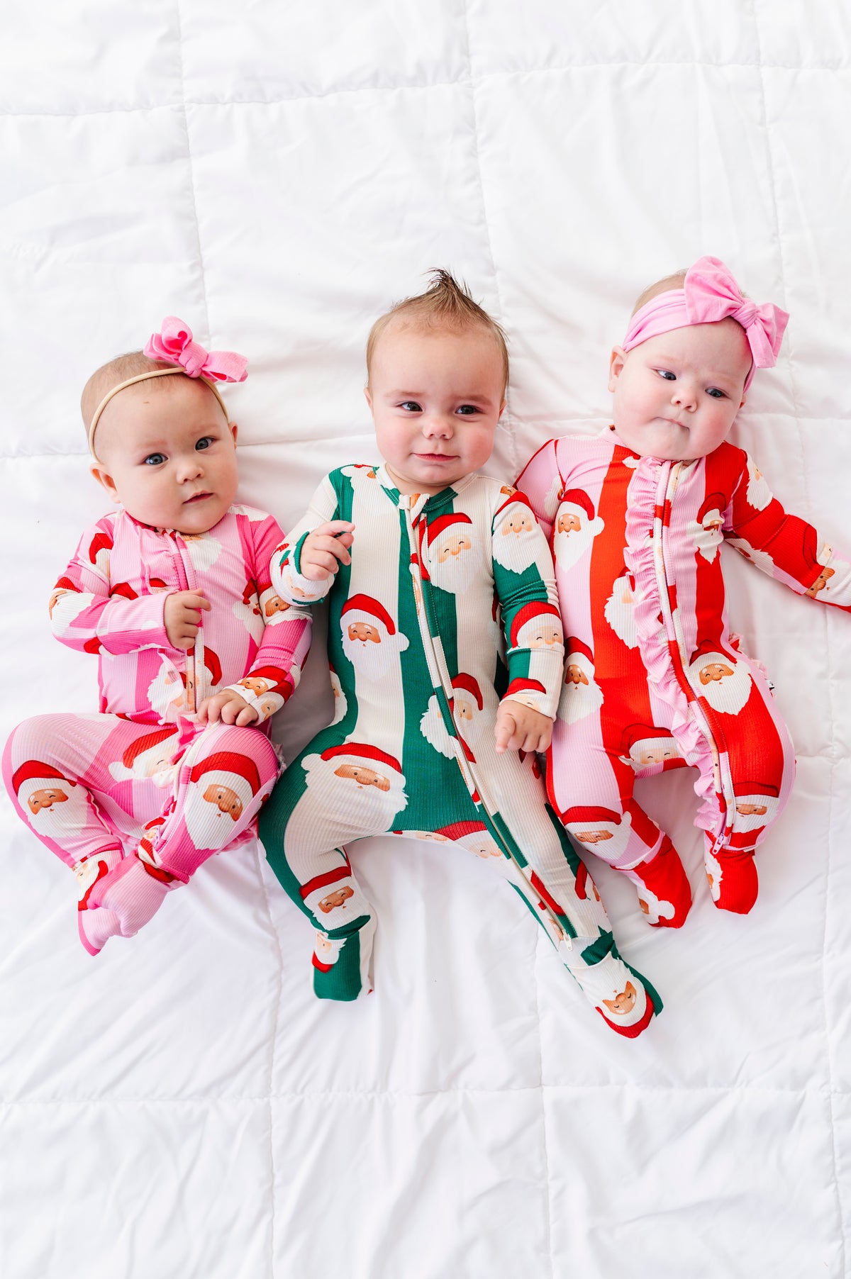Three babies lying side by side on a white quilted blanket, wearing matching Santa Claus print footed bamboo pajamas in pink, green, and red with pink bows in their hair.