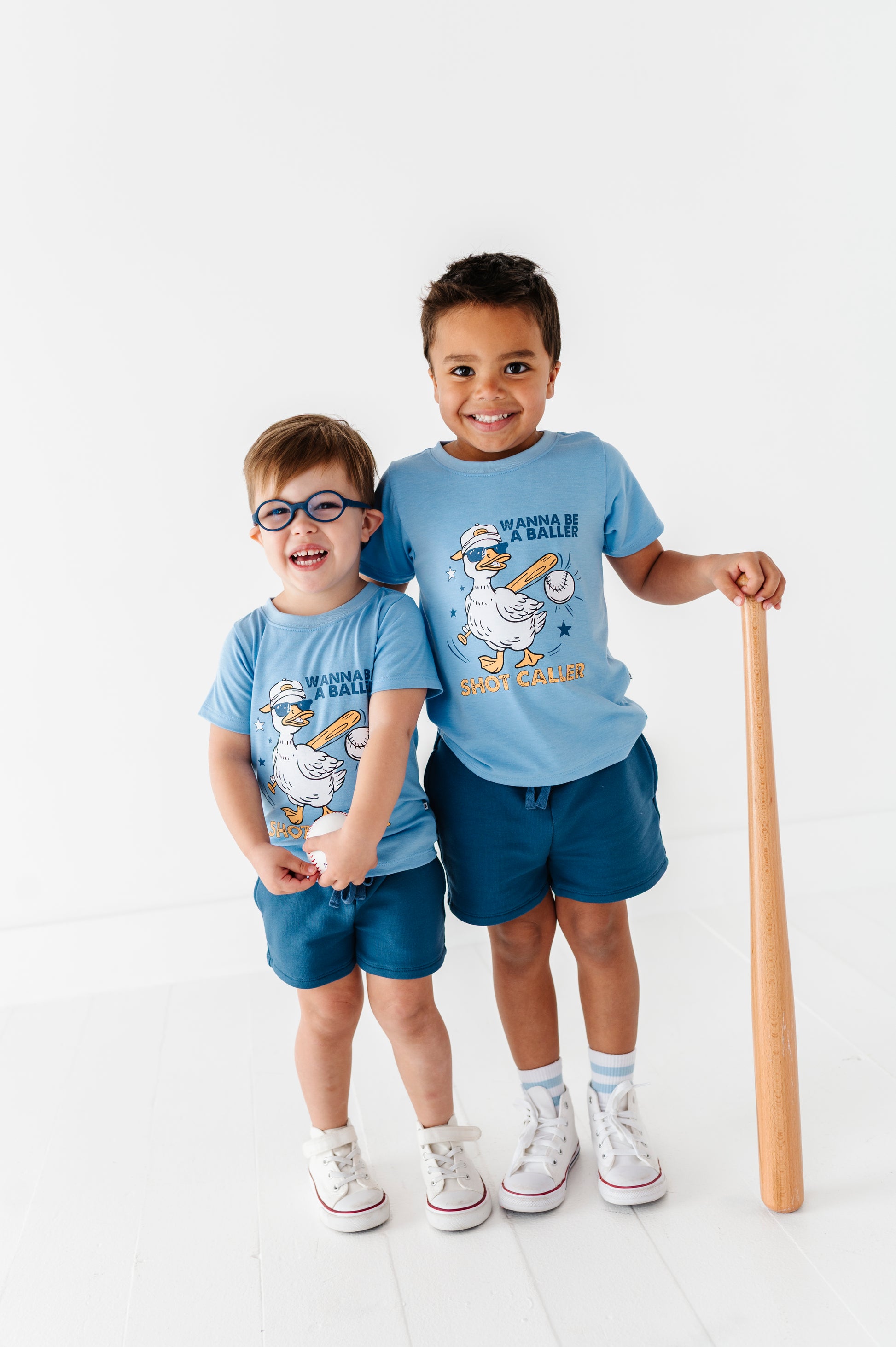 Two children wearing matching blue baseball-themed t-shirts with a cartoon character and holding a baseball bat on a white background.