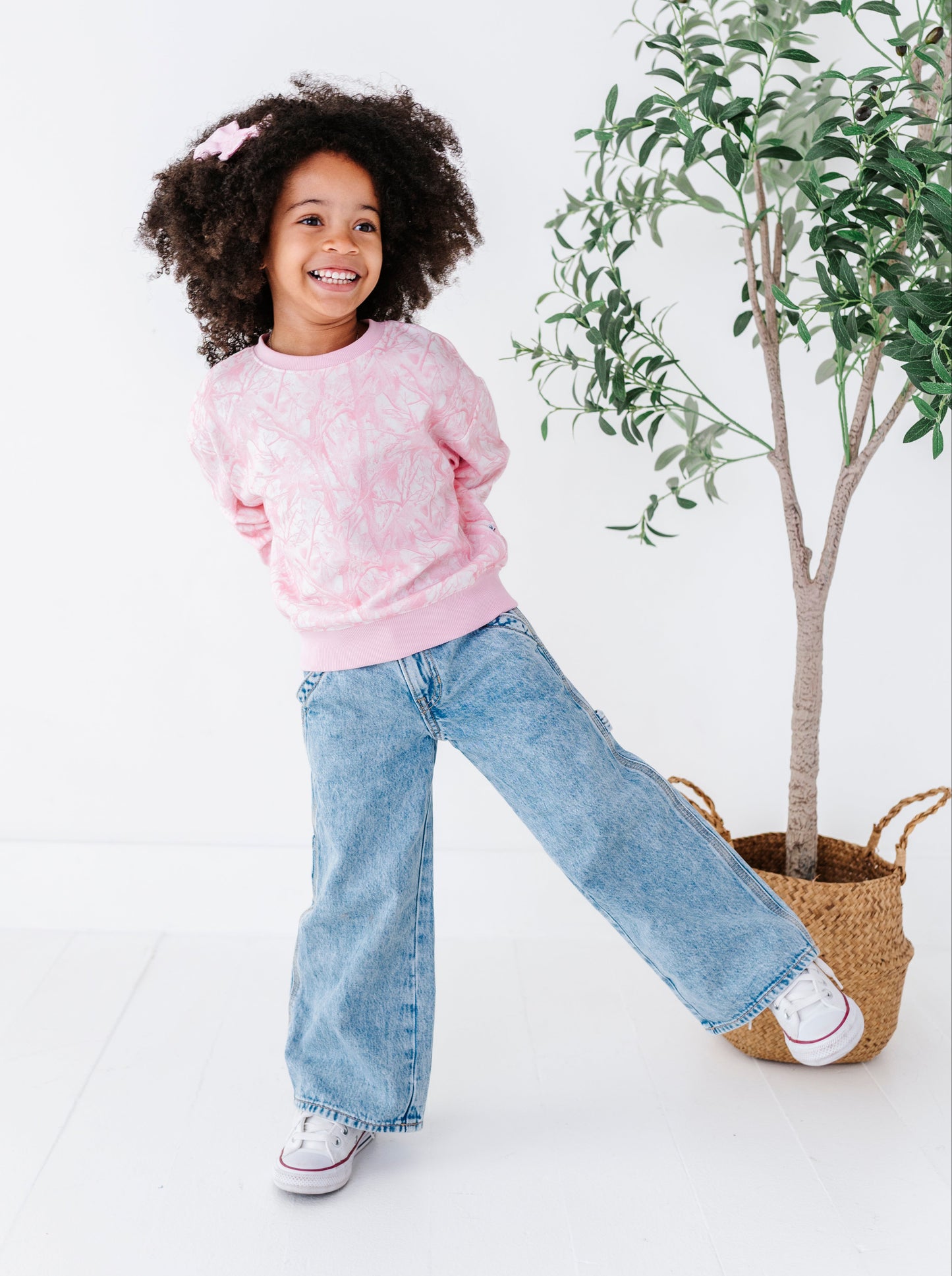 Child wearing a pink camo sweater and blue jeans standing next to a plant on a white background