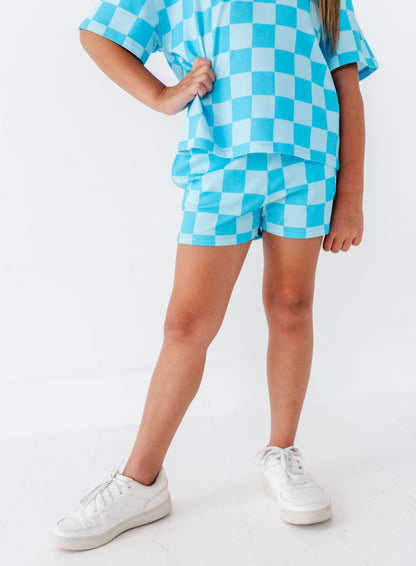 A child wearing a matching light blue and white checkered short-sleeve top and shorts set, paired with white sneakers, standing against a clean white studio background.