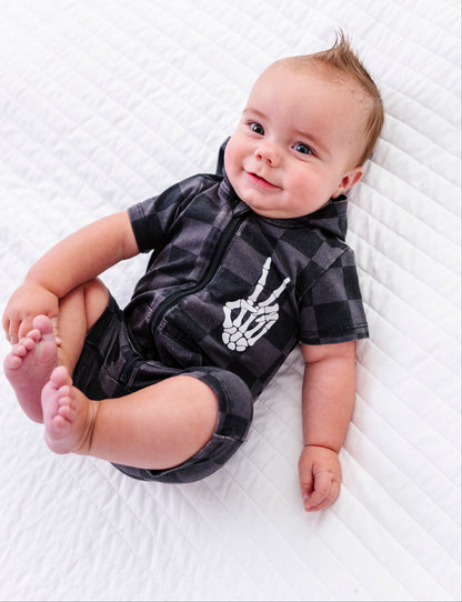 A baby lying on a white quilted blanket, wearing a black and gray plaid short-sleeve romper with a white skeleton hand graphic on the chest.