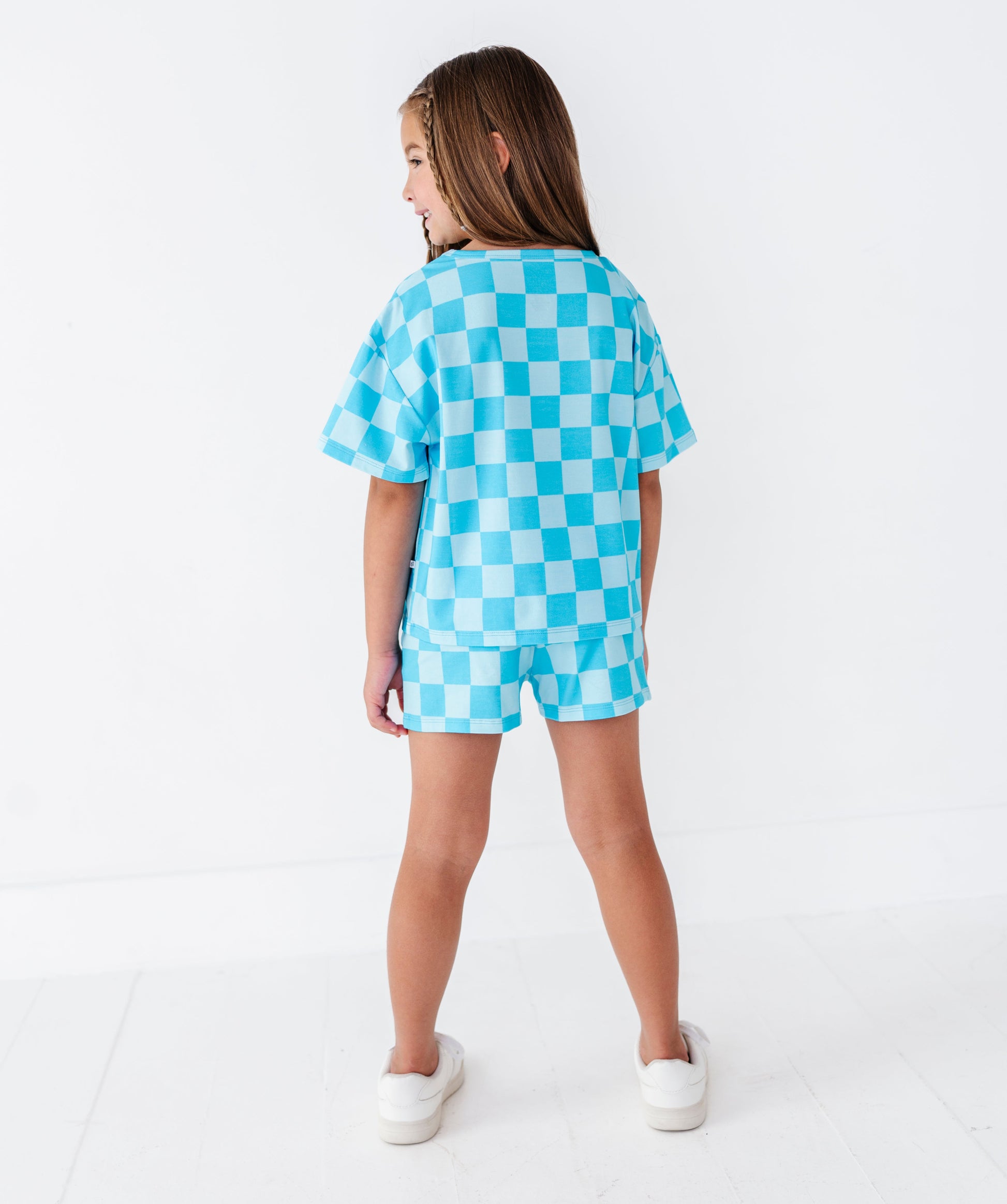 A young girl is shown from behind wearing a light blue and white checkered short-sleeve t-shirt and matching shorts, paired with white slip-on shoes, against a clean white studio background.