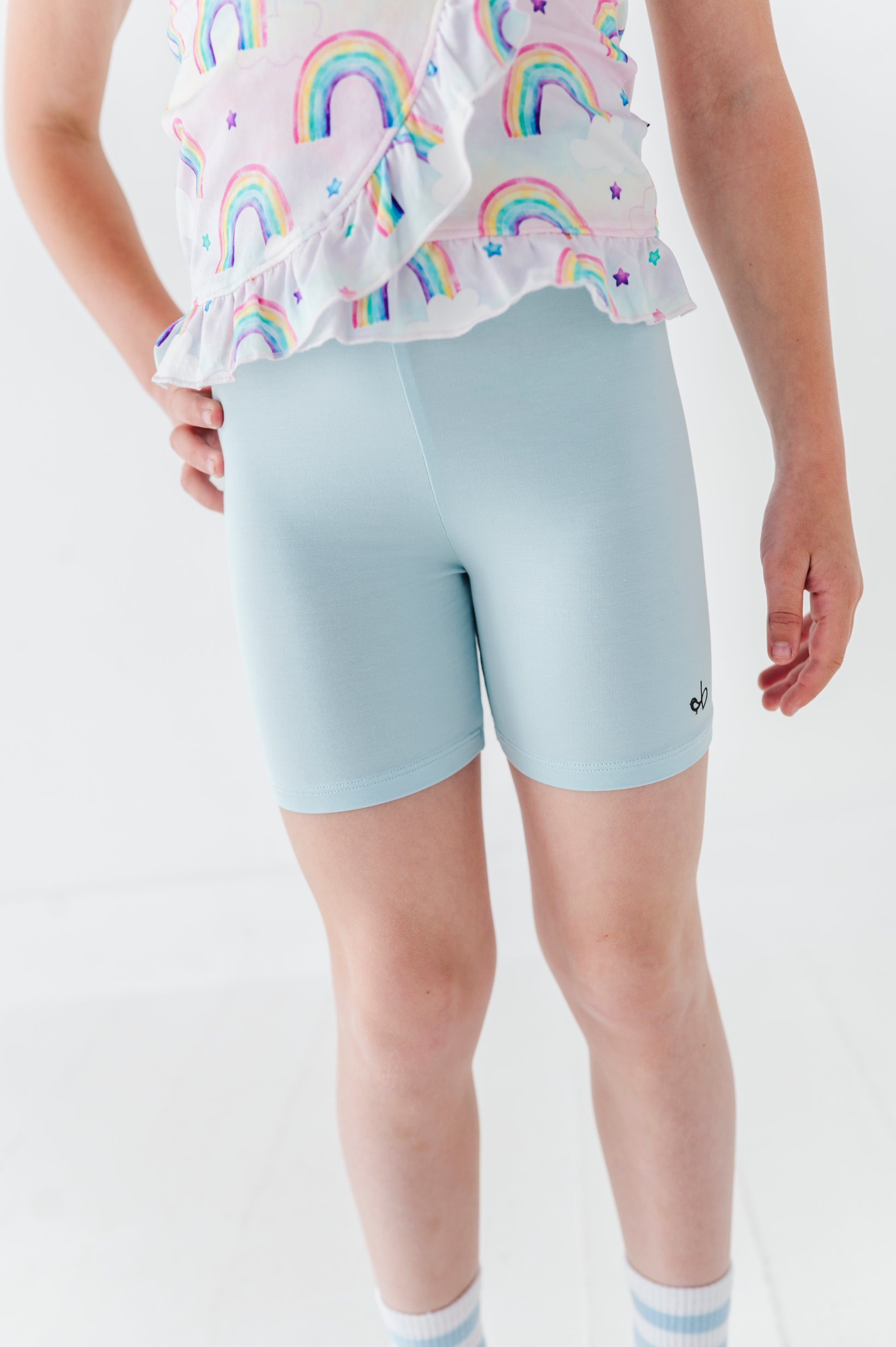 Little girl wearing a bamboo cotton peplum top with watercolor rainbow patterns and light blue bamboo biker shorts on a plain background